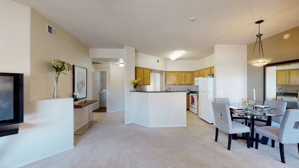 Modern living room here at Dobson Towne Centre Apartments with beige walls and carpet, open to a kitchen with wooden cabinets. A glass dining table with gray chairs sits under a pendant light.