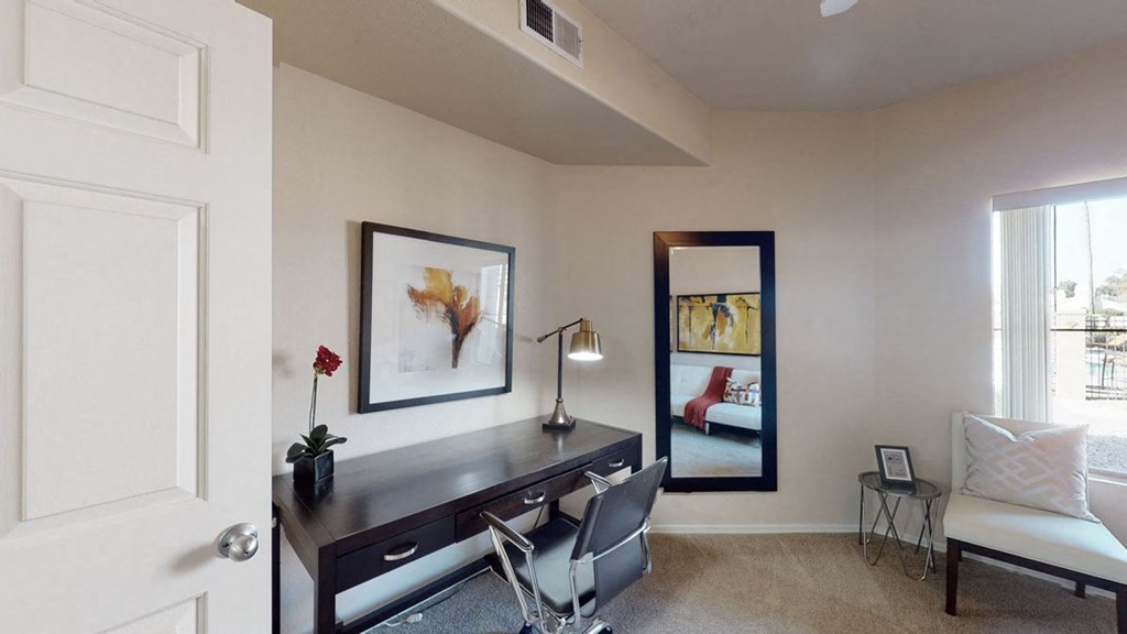 A modern office space here at Dobson Towne Centre Apartments with beige walls features a dark wood desk, a sleek lamp, and a reflective mirror. There's a gray chair, abstract art, and a small plant on the desk, creating a calm and organized atmosphere.