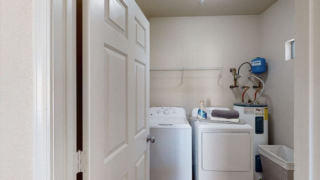 A small laundry room here at Dobson Towne Centre Apartments with a washing machine and dryer. A shelf above holds a detergent bottle and folded towels. A water heater is to the right.