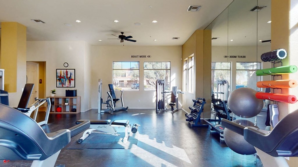 Bright gym here at Dobson Towne Centre Apartments with various equipment, including stationary bikes, dumbbells, yoga mats, and an exercise ball. "BEAST MODE ON" sign adds motivation. Calm and inviting atmosphere.