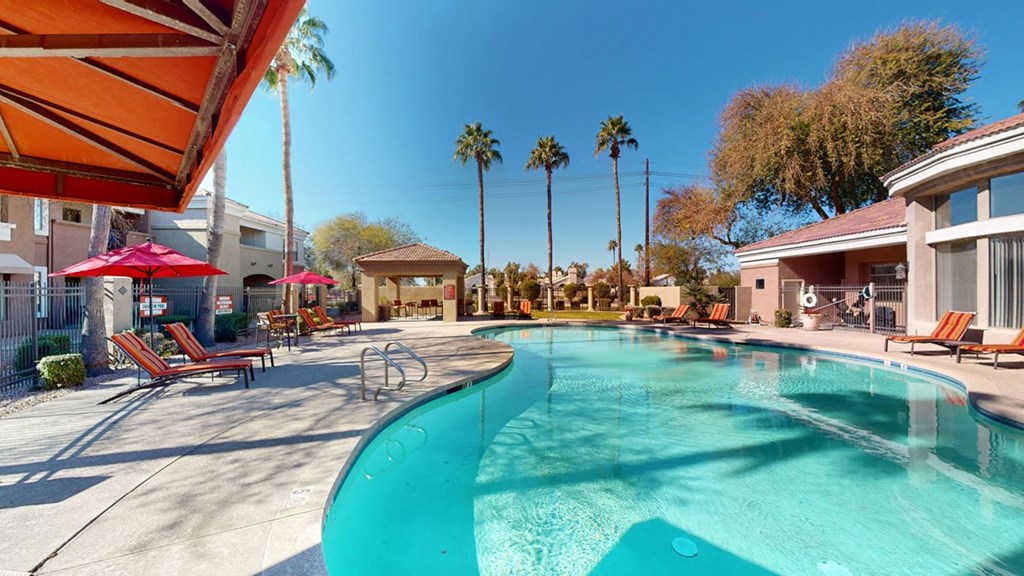 A sunny pool area here at Dobson Towne Centre Apartments with clear blue water, surrounded by lounge chairs under red umbrellas. Palm trees line the background against a clear blue sky.