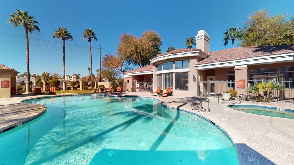 Outdoor pool area here at Dobson Towne Centre Apartments with clear blue water, surrounded by palm trees and sun loungers. Adjacent is a beige building with large windows under a sunny blue sky.