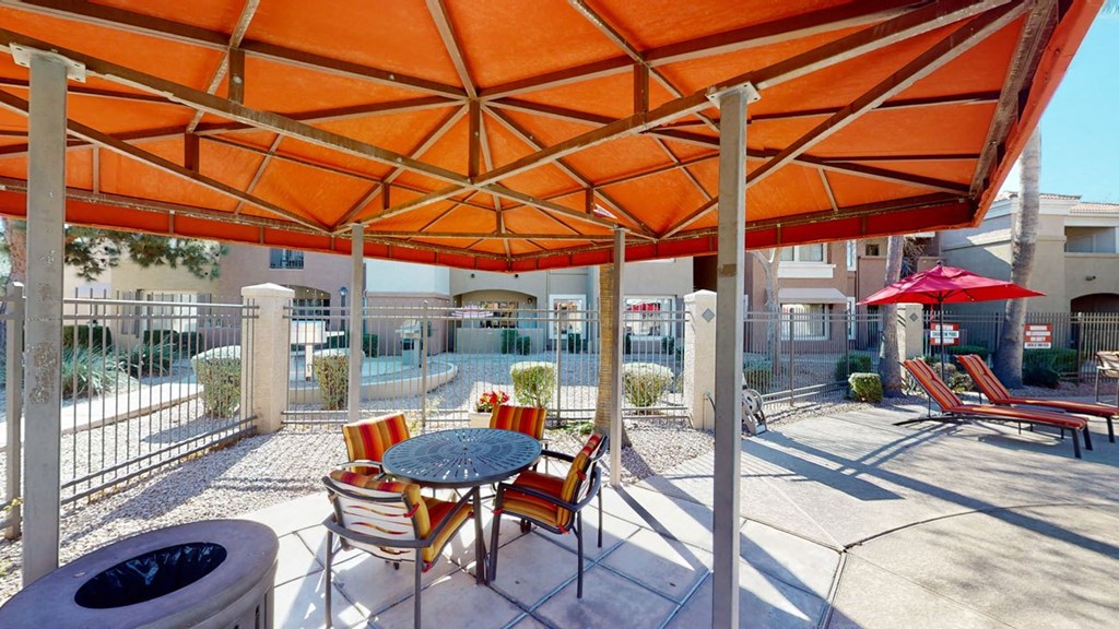 Outdoor patio here at Dobson Towne Centre Apartments under an orange canopy with a round table and striped chairs. Nearby, pool loungers with red umbrellas. Sunny and inviting ambiance.