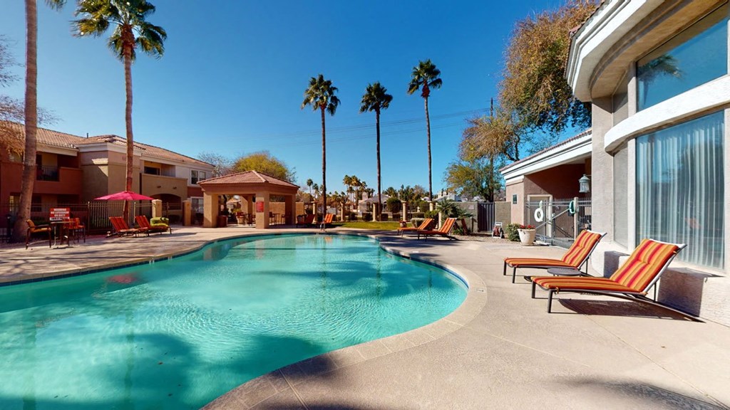 Sunny outdoor pool area here at Dobson Towne Centre Apartments with clear blue water, surrounded by palm trees. Red-striped lounge chairs line the pool deck, creating a relaxing atmosphere.