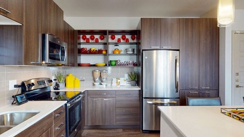 Modern kitchen here at Escape Apartments with dark wood cabinets, stainless steel appliances, and a light countertop. Open shelves display colorful dishes and plants, creating a welcoming and vibrant atmosphere.