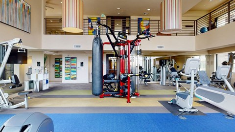 Spacious gym interior with modern equipment here at Escape Apartments, including a red multi-station machine and a punching bag. Brightly colored abstract art decorates the walls.