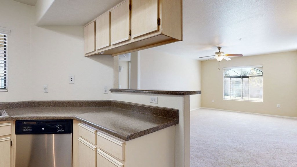 Modern kitchen here at Reserve at Arrowhead Apartments with beige cabinets and speckled countertops opens to a bright living room. Ceiling fan, large window with blinds, and neutral tones.