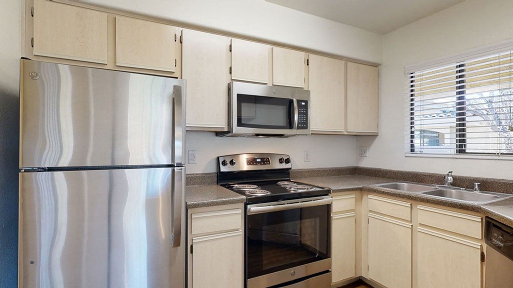 Small kitchen here at Reserve at Arrowhead Apartments with stainless steel appliances, light wood cabinets, and brown countertops. There's a window above the double sink, giving a cozy feel.
