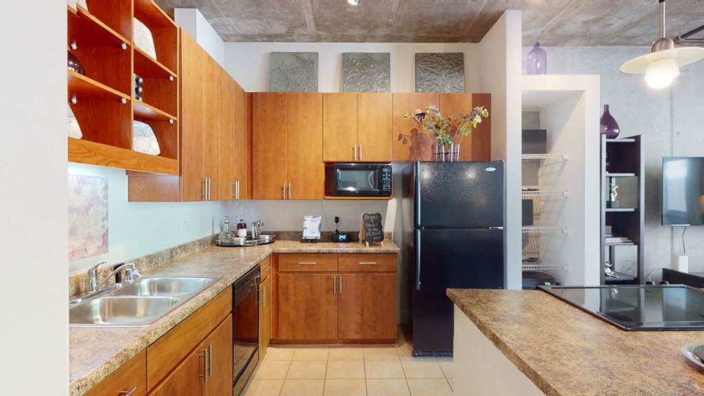Modern kitchen here at Skyline Lofts Apartments with wood cabinets, black appliances, and a granite countertop. A vase with flowers and decorative items add warmth and style.