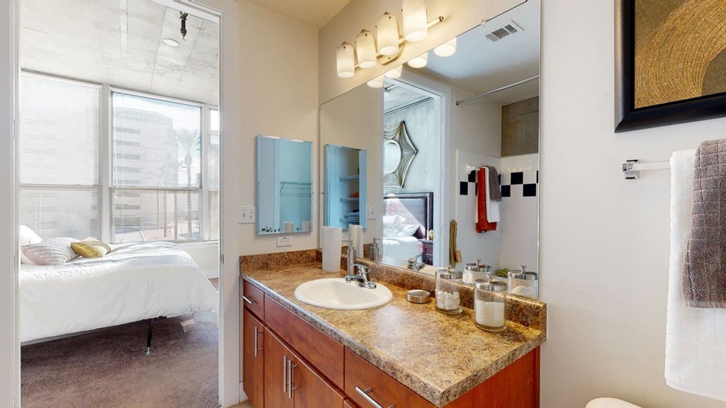 Modern bathroom and bedroom view here at Skyline Lofts Apartments. Marble counter with sink, mirror, and lights. Bedroom has a large window, bed with pillows, and soft lighting.
