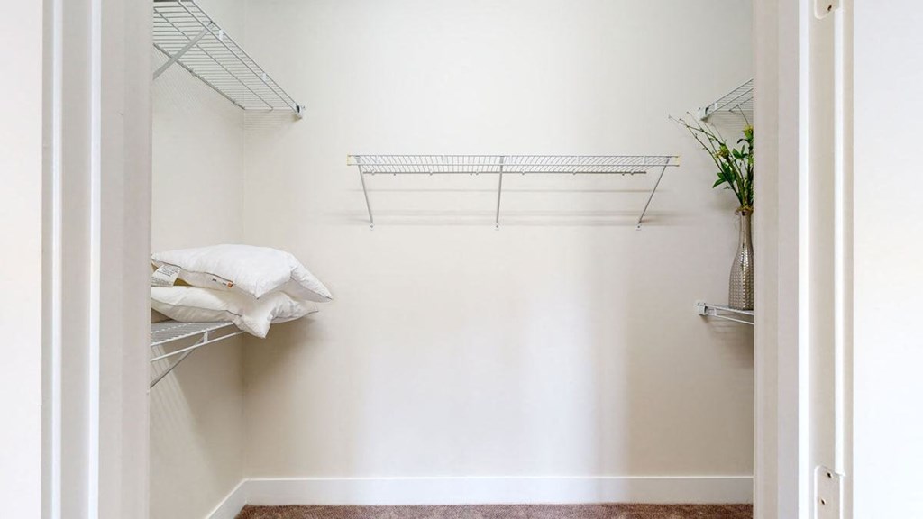 An empty white closet here at Skyline Lofts Apartments with metal shelving on left and back walls. Two pillows sit on the left shelf, and a vase with green plants is on the right shelf.