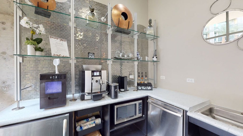 A modern coffee station here at Skyline Lofts Apartments with a silver coffee machine, syrup bottles, and a sign reading "But First Coffee." Shelves hold decorative items and plants.