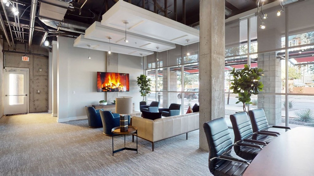 Modern office lounge here at Skyline Lofts Apartments with a cozy atmosphere. Features a digital fireplace on a wall-mounted TV, beige sofa, blue chairs, large conference table, and greenery.