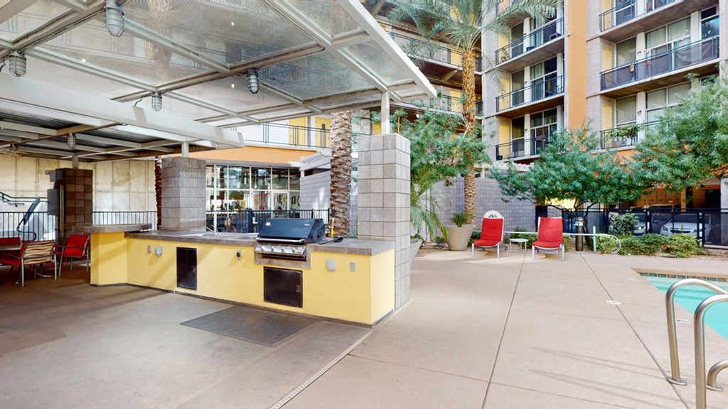 Outdoor patio here at Skyline Lofts Apartments with a covered grilling area, yellow counter and stainless steel BBQ. Nearby, red lounge chairs face a modern apartment building. Relaxed vibe.