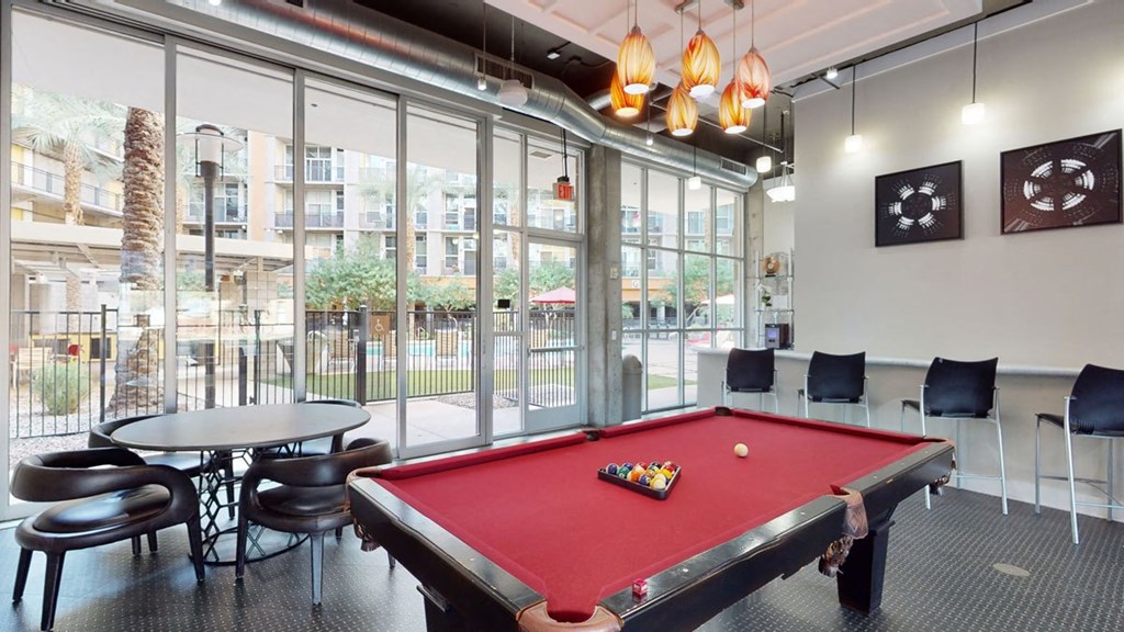 Modern game room here at Skyline Lofts Apartments with a red pool table, four artistic pendant lights, and a round table with chairs. Large windows show a pool area outside.
