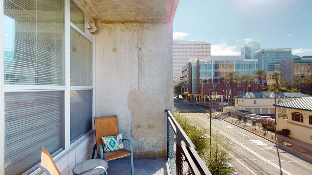 A small apartment balcony here at Skyline Lofts Apartments with two brown chairs, one with a colorful cushion, and a round table. Overlooks a sunny urban street lined with palm trees and modern buildings.