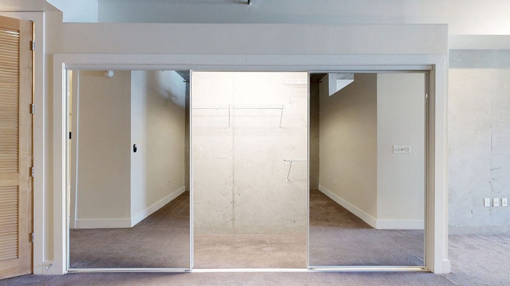 Spacious, empty walk-in closet here at Skyline Lofts Apartments with mirrored sliding doors, light beige walls, and brown carpet, creating a clean and minimalistic feel.