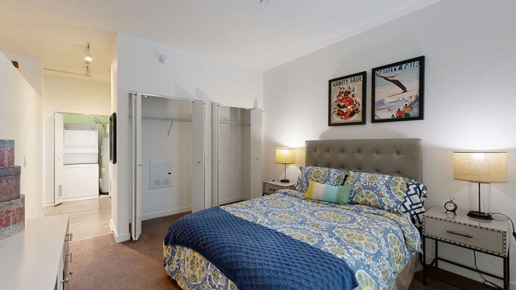 Cozy bedroom here at Trend at 51 Apartments with a patterned bedspread, two bedside lamps, and framed art. Open closet and laundry area visible. Warm, inviting atmosphere.