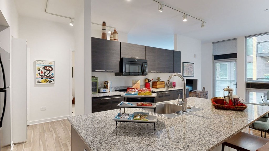 Modern kitchen here at Trend at 51 Apartments with granite countertops, dark cabinets, and a central island. It features a sink, snacks on display, and a cozy, well-lit ambiance.