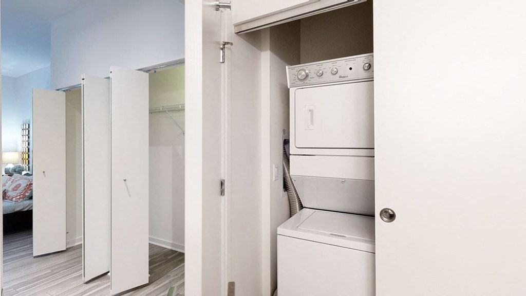 Compact laundry area here at Trend at 51 Apartments with a stacked washer and dryer tucked in a closet with sliding doors. Adjacent is an open closet with white bi-fold doors.