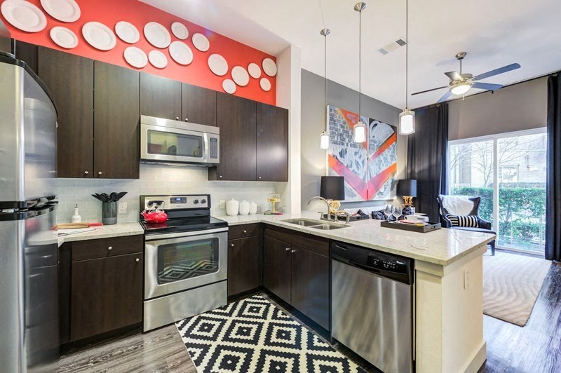 Modern kitchen here at Arpeggio Apartment Homes with dark wood cabinets and stainless steel appliances. Red accent wall with white plates. Geometric rug. Living area with large window. Cozy and stylish.