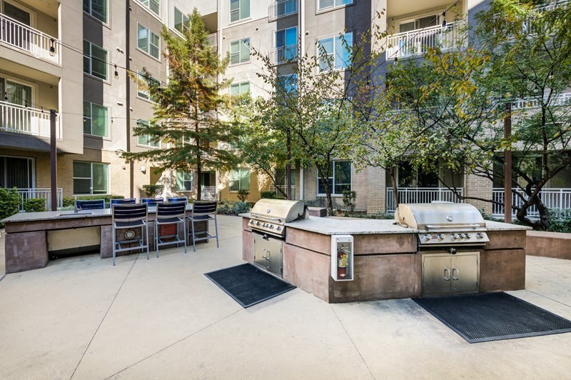Outdoor communal grilling area here at Arpeggio Apartment Homes with two stainless steel barbecue grills on a stone island. Surrounded by apartment buildings, trees, and seating.