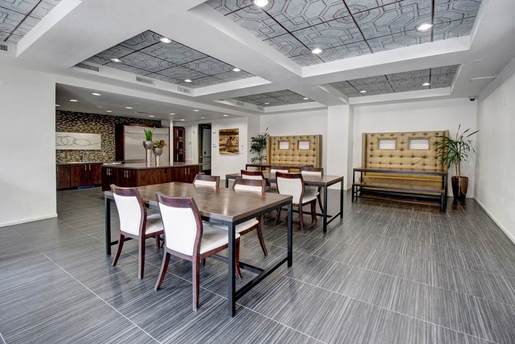 Modern dining area here at Artisan at Main Street Metro Apartments with gray tiled floor, wooden table, and white cushioned chairs. Stylish tufted benches and potted plants add a cozy feel.