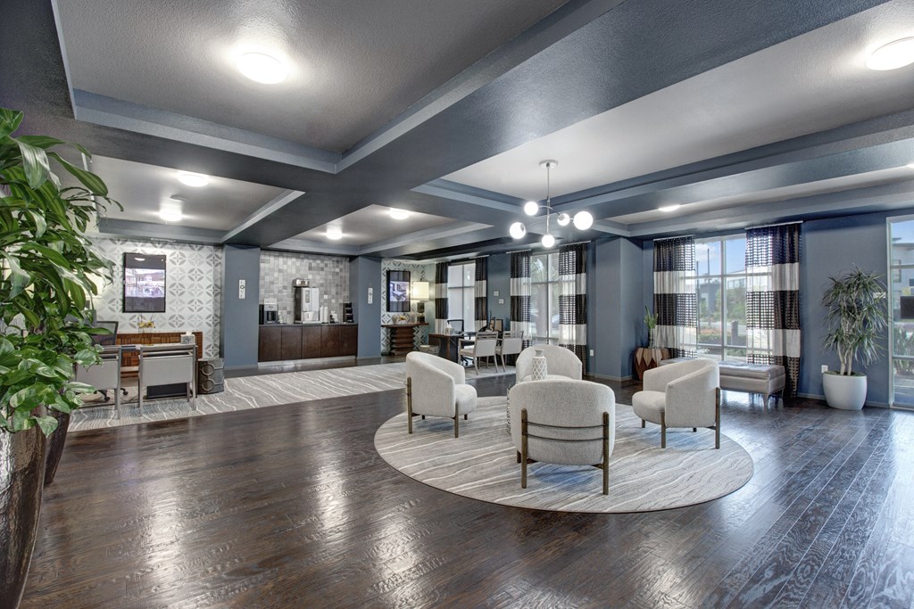 Modern lounge here at Artisan at Main Street Metro Apartments with dark wood floors, round rug, and plush chairs. A coffee station and tables with chairs are along the patterned walls. Bright, airy atmosphere.