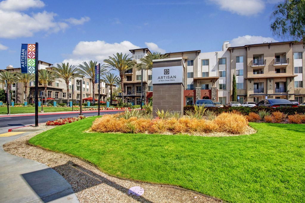 Modern apartment complex with lush green lawn in foreground. Sign reads “Artisan at Main Street Metro.” Palm trees and colorful banners line the street, under a blue sky.