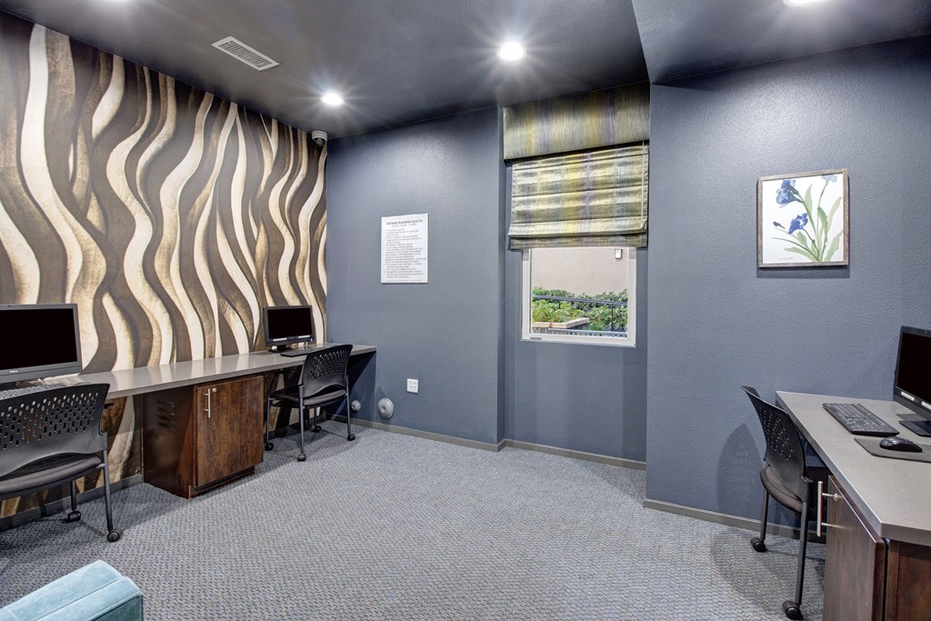 Modern computer room here at Artisan at Main Street Metro Apartments with a wavy patterned wall, two desks with computers, and chairs. A window with a striped blind and framed plant artwork add color.
