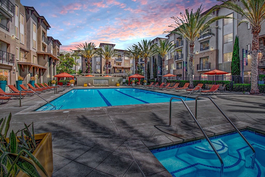 Modern Artisan at Main Street Metro apartment complex at sunset with a central pool, surrounded by palm trees and lounge chairs. The sky is vibrant with pink and blue hues, creating a relaxing ambiance.