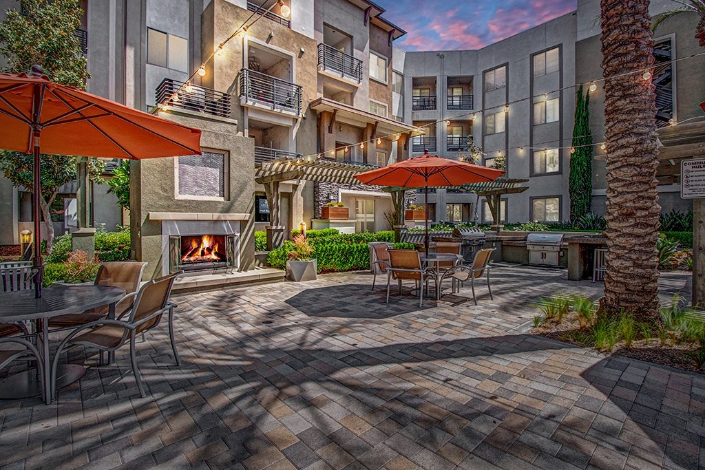 Cozy outdoor patio here at Artisan at Main Street Metro Apartments with tables, orange umbrellas, and string lights. A lit fireplace adds warmth. Surrounded by modern apartments at dusk.