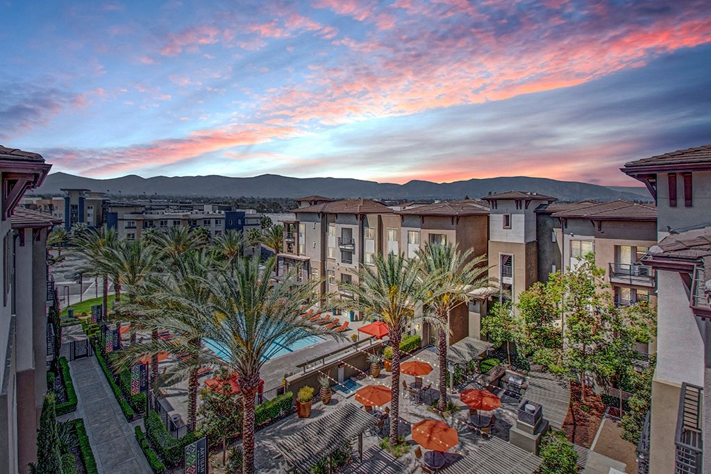A serene Artisan at Main Street Metro apartment complex with palm trees, a pool, and orange umbrellas on a patio. The sky is vibrant with pink and blue hues, creating a tranquil mood.