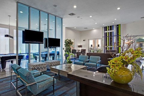 Modern lounge area here at Aspire at Pinnacle Peak Apartments with blue chairs, patterned ottomans, glass divider, and large TVs. Scenic decor includes green plants and a bright, spacious interior.