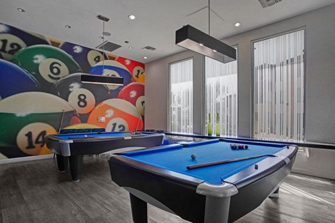 Modern billiards room here at Aspire at Pinnacle Peak Apartments with two blue pool tables, cue sticks, and balls. Features a colorful mural of large billiard balls and tall windows.
