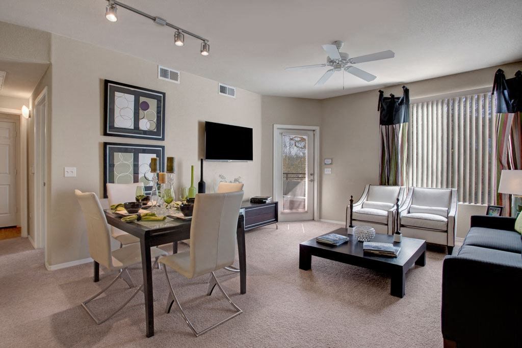 Modern living and dining space here at Aspire at Pinnacle Peak Apartments with neutral tones. A glass dining table with white chairs, abstract art, a wall-mounted TV, and cozy seating with natural light.