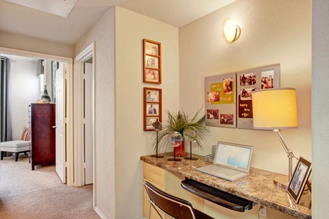 A cozy home office space here at Aspire at Pinnacle Peak Apartments with a marble desk, open laptop, modern chair, and a warm lamp. Family photos adorn the wall, creating a welcoming ambiance.