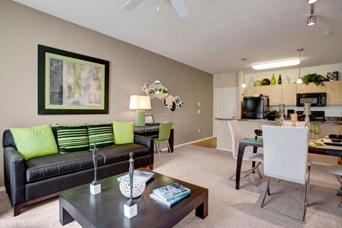 Modern living room here at Aspire at Pinnacle Peak Apartments with black sofa and green cushions, abstract art, and a sleek kitchen. Warm lighting and neutral tones create a cozy ambiance.