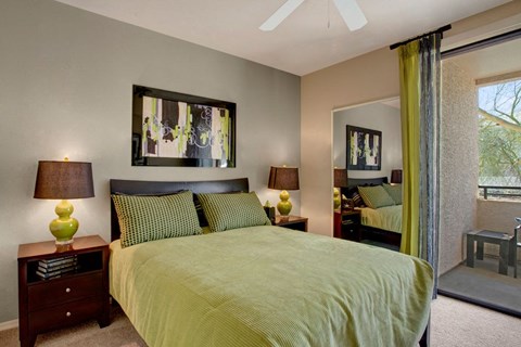 Modern bedroom here at Aspire at Pinnacle Peak Apartments with green-themed decor. Features include a queen bed, abstract artwork, matching lamps, wooden side tables, and a glass door leading to a balcony.