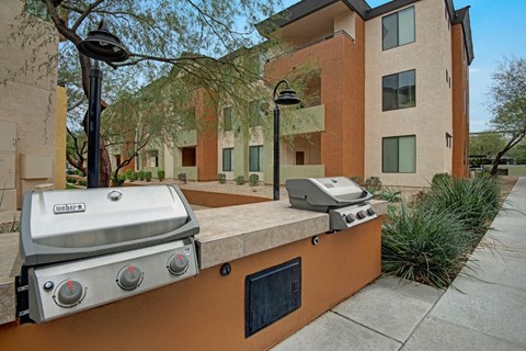 Two stainless steel barbecue grills here at Aspire at Pinnacle Peak Apartments are on a stone countertop in a modern apartment complex courtyard, surrounded by green plants and walking paths.