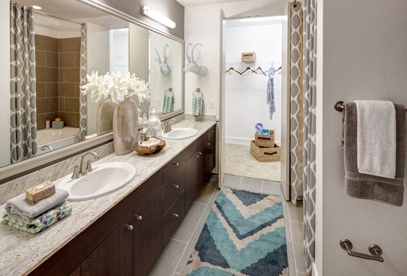 Modern bathroom here at Arpeggio Apartment Homes with dual sinks, marble countertop, and decorative items. Blue zigzag rug leads to a walk-in closet. Neutral tones, elegant vibe.