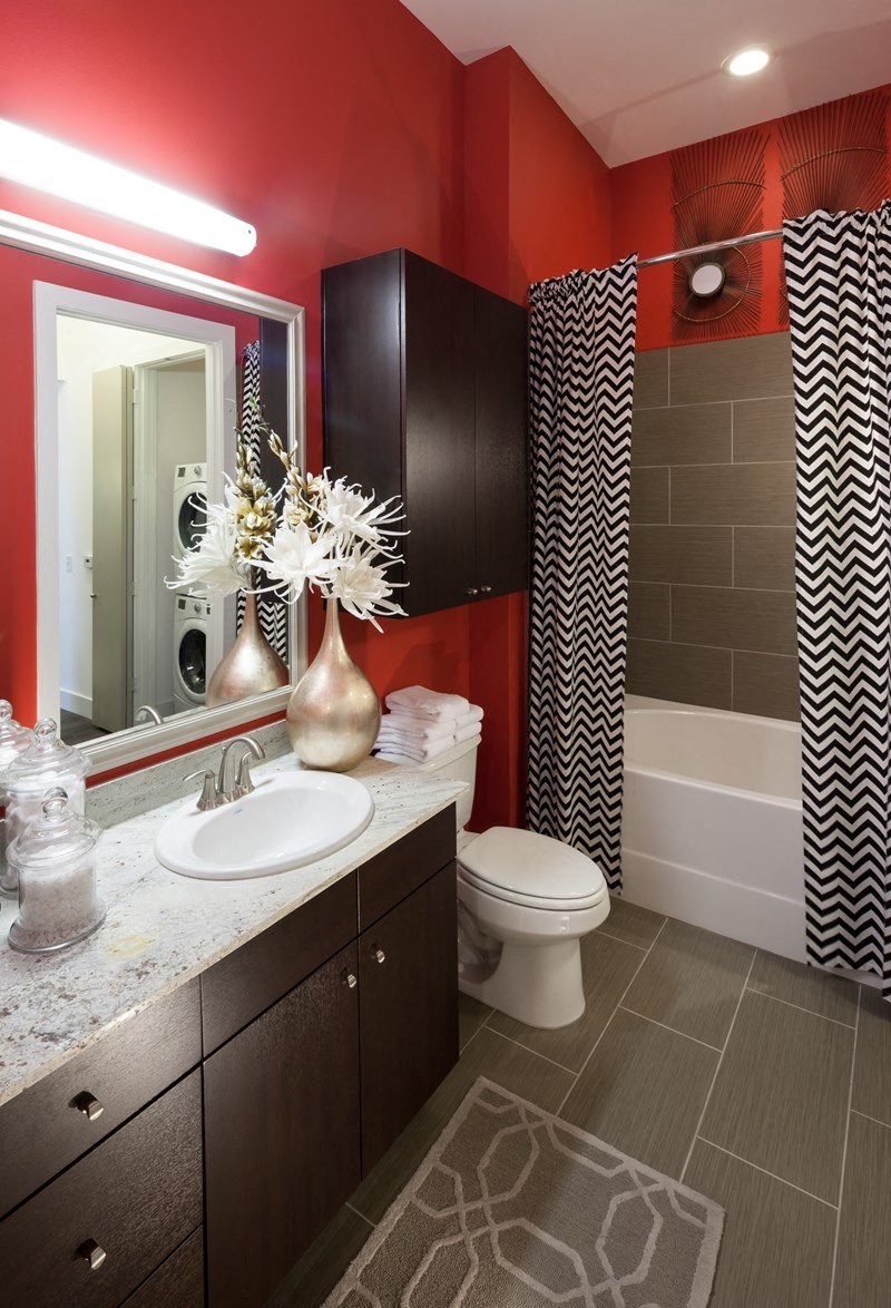 Modern bathroom here at Arpeggio Apartment Homes with bold red walls, dark wood cabinets, and zigzag-patterned shower curtain. Features a white sink, large mirror, and elegant decor.