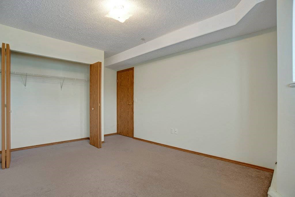 Empty bedroom here at Aspen Terrace Apartment Homes with a beige carpet, white walls, and a ceiling light. Open closet with wooden doors and a shelf. Quiet and minimalistic tone.