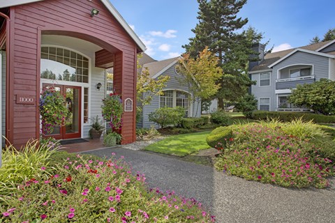 A red house with a white door and windows is surrounded by a garden with flowers and trees.