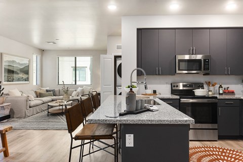 Modern open-plan kitchen and living room here at Reflect at Dobson Ranch Apartments. Gray kitchen cabinets and granite island with brown stools. Cozy cream sofa in lounge area with large windows.