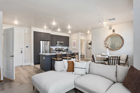 Modern open-plan living area here at Reflect at Dobson Ranch Apartments with a cozy beige sectional sofa, a sleek kitchen with dark cabinets, a round dining table, and warm lighting.