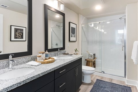 Modern bathroom here at Reflect at Dobson Ranch Apartments with two sinks on a granite countertop, large mirrors, and a glass-enclosed shower. Decor includes framed art and subtle plants.