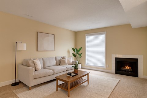 A cozy living room here at 3600 Brentwood with soft beige carpet, a light gray sofa, a wooden coffee table, and a warm fireplace, accented by a bright window with white blinds and simple modern decor.