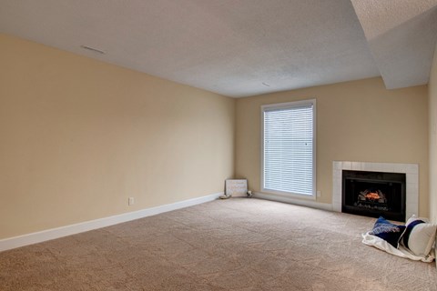 A spacious, empty living room here at 3600 Brentwood with soft beige carpet, a bright window with white blinds, and a cozy fireplace that adds warmth to the open space.