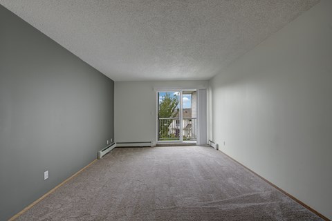 A spacious, empty living room here at Aspen Terrace with soft carpet, neutral walls, and a sliding glass door that opens to a private balcony. A large window and bright natural light create an airy, inviting feel.